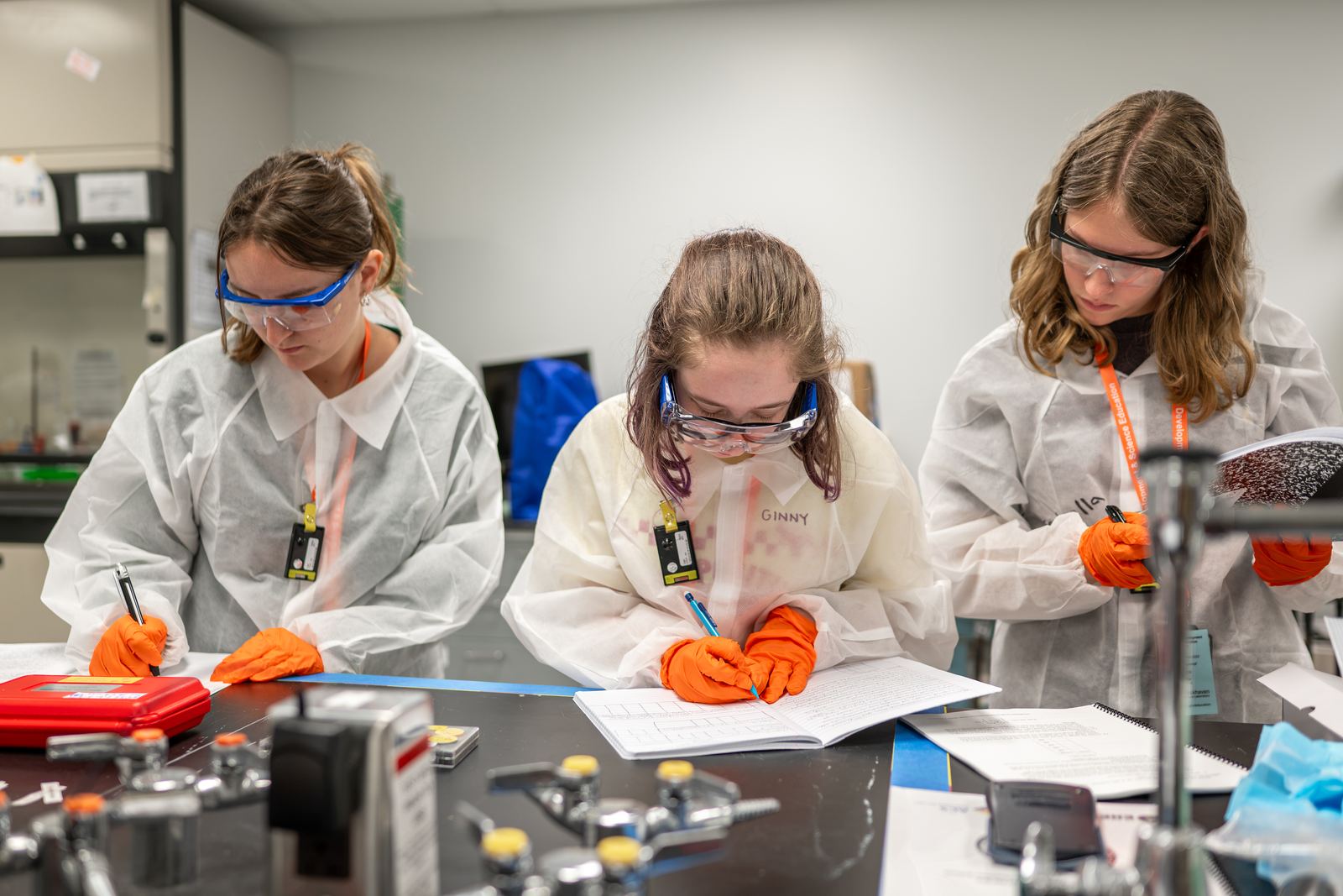 Three NCSS students in lab coats, safety glasses and gloves record results in lab notebooks